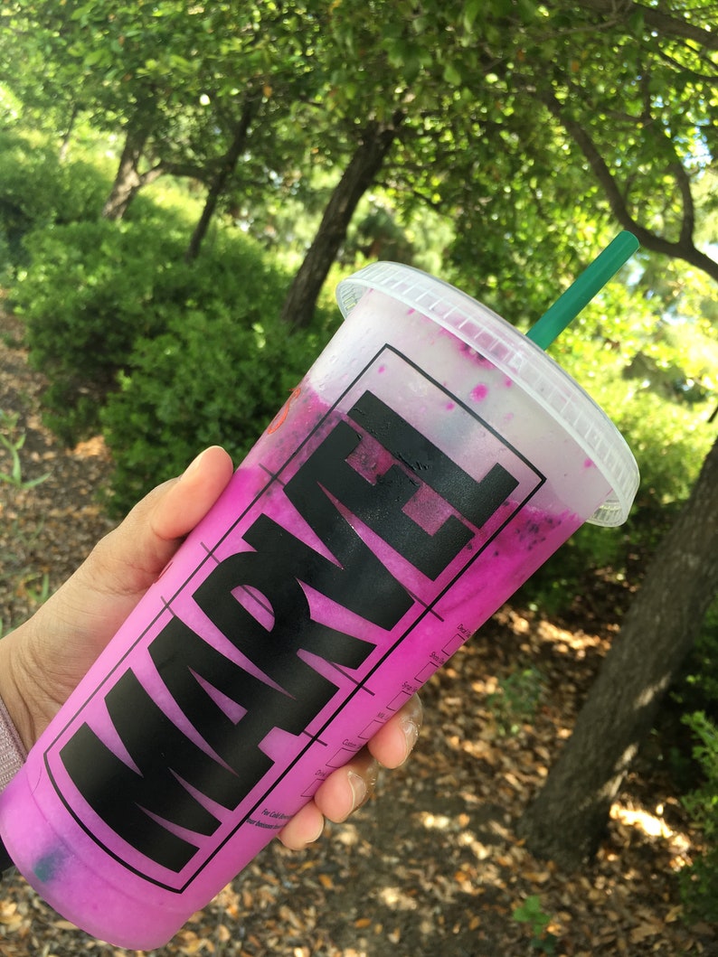 Puede incluir: Un vaso de pl&aacute;stico rosa con un logotipo MARVEL negro y una pajita verde. El vaso est&aacute; lleno de una bebida rosa y morada.