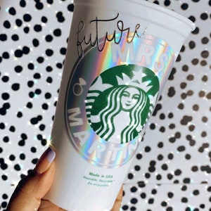 Op de afbeelding: Witte herbruikbare beker met een holografische sticker waarop "future mrs" staat en een groen Starbucks-logo. De sticker heeft een regenboogkleurig, iriserend effect.