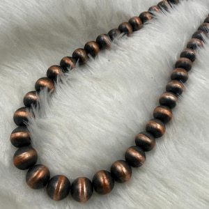 May include: A necklace composed of spherical beads featuring a metallic copper finish with darkened, oxidized accents. The beads are uniform in size and strung together in a continuous strand, displaying a reflective sheen and a textured appearance.