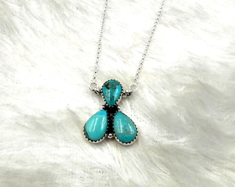 May include: A silver necklace featuring a pendant with four turquoise stones. The pendant has a central teardrop-shaped stone and three larger, pear-shaped stones. The necklace is displayed on a white, textured surface.