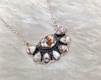 May include: A silver necklace featuring a pendant with a central oval stone surrounded by smaller, teardrop-shaped stones. The stones have a mix of white and brown patterns. The necklace chain is delicate and silver.