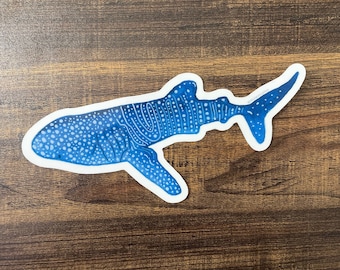 May include: A blue and white whale shark sticker with a spotted pattern. The sticker is on a brown wooden surface.