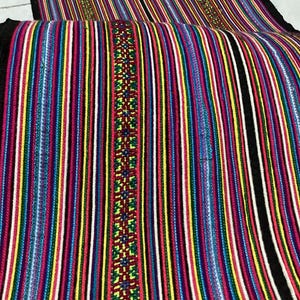 May include: A colorful woven textile with vertical stripes in various colors including red, yellow, blue, and white. A central band features a detailed geometric pattern. The edges are finished with a black border. This textile is likely used for decorative purposes.