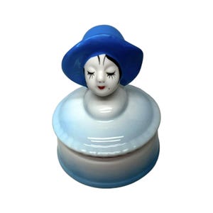 May include: A ceramic trinket box depicting a woman's head with a blue hat. The face has closed eyes and red lips. The box is white and light blue, with a lid that resembles the woman's dress. A decorative item.