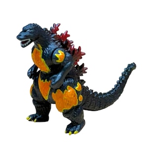 May include: A black Godzilla toy with orange and yellow accents on its chest and legs. The toy has red dorsal fins and yellow claws. The Godzilla figure is standing with its mouth open, revealing white teeth.