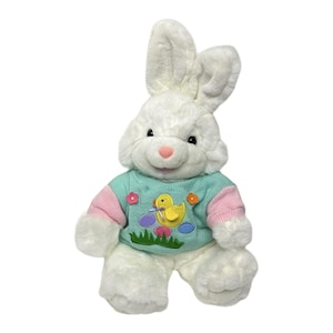 May include: A white plush Easter bunny wearing a light blue sweater with pink sleeves. The sweater features a yellow duck, colorful eggs, and flowers. The bunny has long ears, a pink nose, and black eyes.