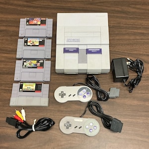May include: A light grey Super Nintendo game console with two matching controllers, a black power adapter, an AV cable, and four grey game cartridges. The console features purple power and reset buttons, and the cartridges include labels for Super Mario World and other titles.