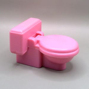 May include: A small, pink toy toilet with a rectangular tank and a rounded seat. The toilet is made of plastic and has a smooth, glossy finish. The seat is closed. The overall design is simple and child-like.