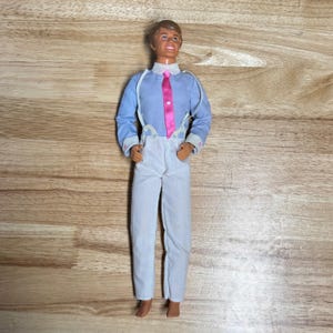 May include: A vintage doll dressed in a light blue shirt, pink tie, and white trousers. The doll has brown hair and white braces. The pink tie features a heart. The doll is standing on a wooden surface.