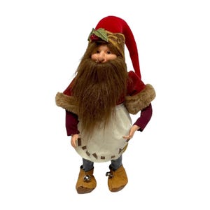 Puede incluir: Una muñeca de gnomo navideño con una larga barba marrón, que lleva un sombrero de terciopelo rojo con detalles de acebo, un abrigo rojo con ribete de piel sintética y un delantal color crema. El gnomo tiene botas marrones con campanas.