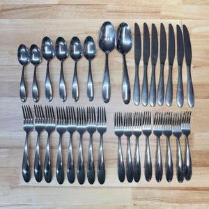 以下が含まれることがあります： 木製の表面に配置されたステンレス製のカトラリーセット。さまざまなサイズのスプーン、フォーク、ナイフが含まれています。光を反射する、洗練されたモダンなデザインです。