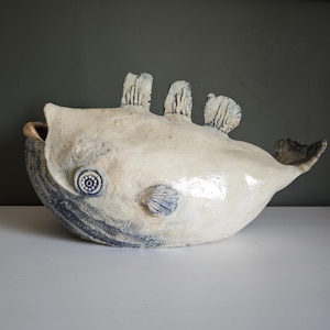 Può includere: Una scultura in ceramica a forma di pesce con un corpo testurizzato color bianco sporco e dettagli blu scuro. Il pesce presenta tre pinne dorsali, un occhio circolare con un centro decorato e linee incise lungo la mascella inferiore.