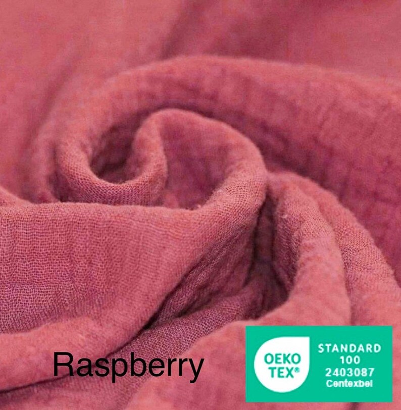 May include: Close-up of raspberry-colored fabric with soft folds and textures. The word "Raspberry" is printed in black at the bottom left. A green and white "OEKO-TEX" label is in the bottom right corner.