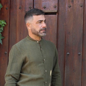Könnte beinhalten: Ein Mann mit kurzen, dunklen Haaren und Bart trägt ein grünes, langärmeliges Hemd mit Stehkragen. Er steht vor einer Holztür.