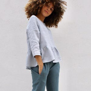 Könnte beinhalten: Ein hellblaues Leinenoberteil mit Rüschensaum und Dreiviertelärmeln wird mit einer teal-farbenen Leinenhose kombiniert. Das Outfit ist schlicht und elegant, perfekt für den Alltag. Das Model hat lockiges braunes Haar.