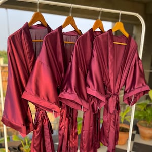 May include: Four burgundy satin robes hanging on a white rack. The robes have long sleeves and a tie belt.