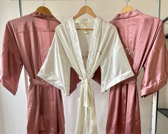 May include: Three satin robes hanging on a white rack. The robes are in shades of pink and white. The white robe has the word "Bride" embroidered on it. The pink robes have the words "Maid of Honor" and "Bridesmaid" embroidered on them.