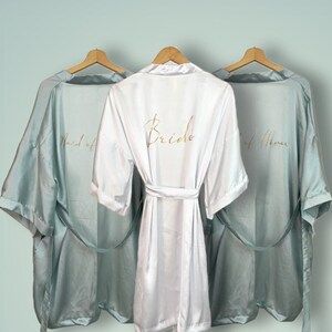 Könnte beinhalten: Drei Satin-Roben hängen an Kleiderbügeln. Die Robe in der Mitte ist weiß mit dem Wort "Bride" in goldener Schrift gestickt. Die Roben auf beiden Seiten sind hellblau mit den Worten "Maid of Honor" und "Best of Honor" in goldener Schrift gestickt.