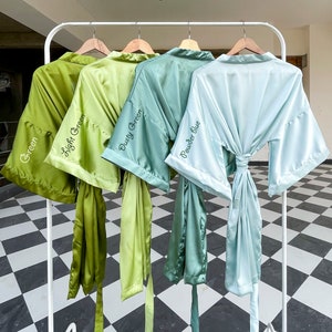 Op de afbeelding: Vier satijnen badjassen hangen aan een witte rek. De badjassen zijn in tinten groen en blauw. De badjassen zijn gelabeld met "Green", "Light Green", "Dusty Green" en "Powder Blue".