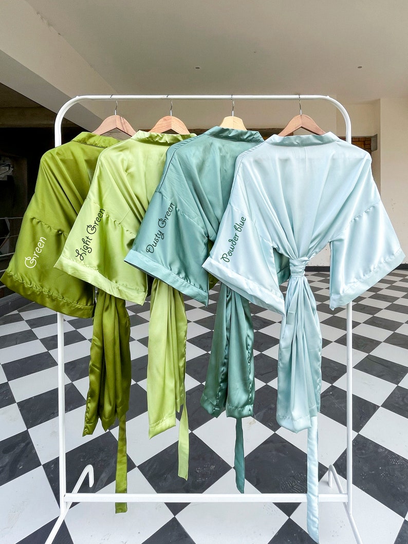 Op de afbeelding: Vier satijnen badjassen hangen aan een witte rek. De badjassen zijn in tinten groen en blauw. De badjassen zijn gelabeld met "Green", "Light Green", "Dusty Green" en "Powder Blue".