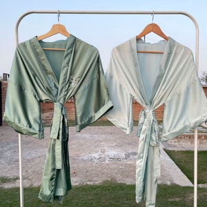 Op de afbeelding: Twee satijnen badjassen hangen aan een rek. De badjas aan de linkerkant is saliegroen, terwijl de badjas aan de rechterkant lichtblauw is. Beide badjassen hebben een strik in de taille en zijn gemaakt van een zijdeachtig materiaal.