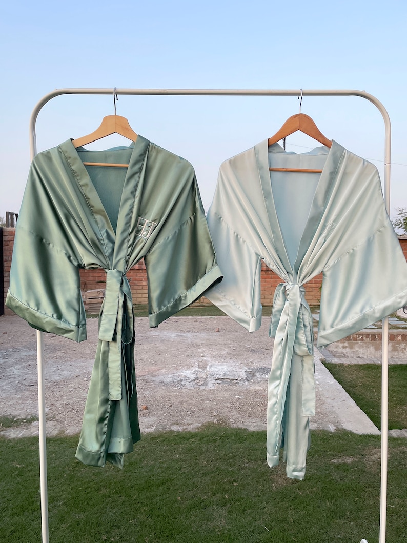 Op de afbeelding: Twee satijnen badjassen hangen aan een rek. De badjas aan de linkerkant is saliegroen, terwijl de badjas aan de rechterkant lichtblauw is. Beide badjassen hebben een strik in de taille en zijn gemaakt van een zijdeachtig materiaal.