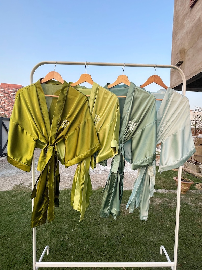 Op de afbeelding: Vier satijnen badjassen in verschillende tinten groen en blauw hangen aan een witte rek. De badjassen hebben een strik in de taille en zijn geborduurd met de letter "R".