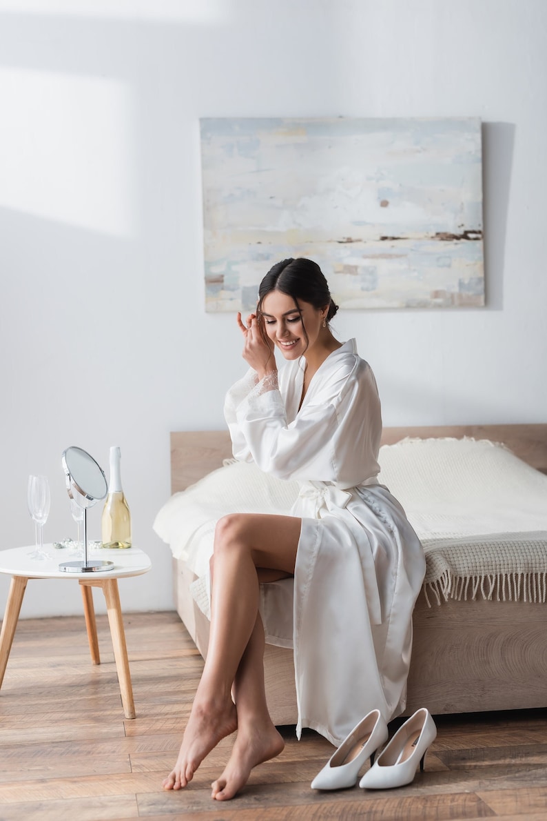 May include: A woman in a white satin robe sits on a bed, smiling as she adjusts her hair. She is wearing white high heels and has a glass of champagne on a side table.