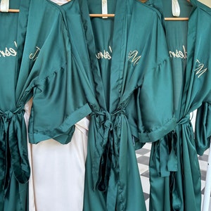 May include: Three green satin robes with gold embroidery. The robes have a sash tie and are hanging on wooden hangers. The embroidery on the robes reads "Bride" and "Bridesmaid".