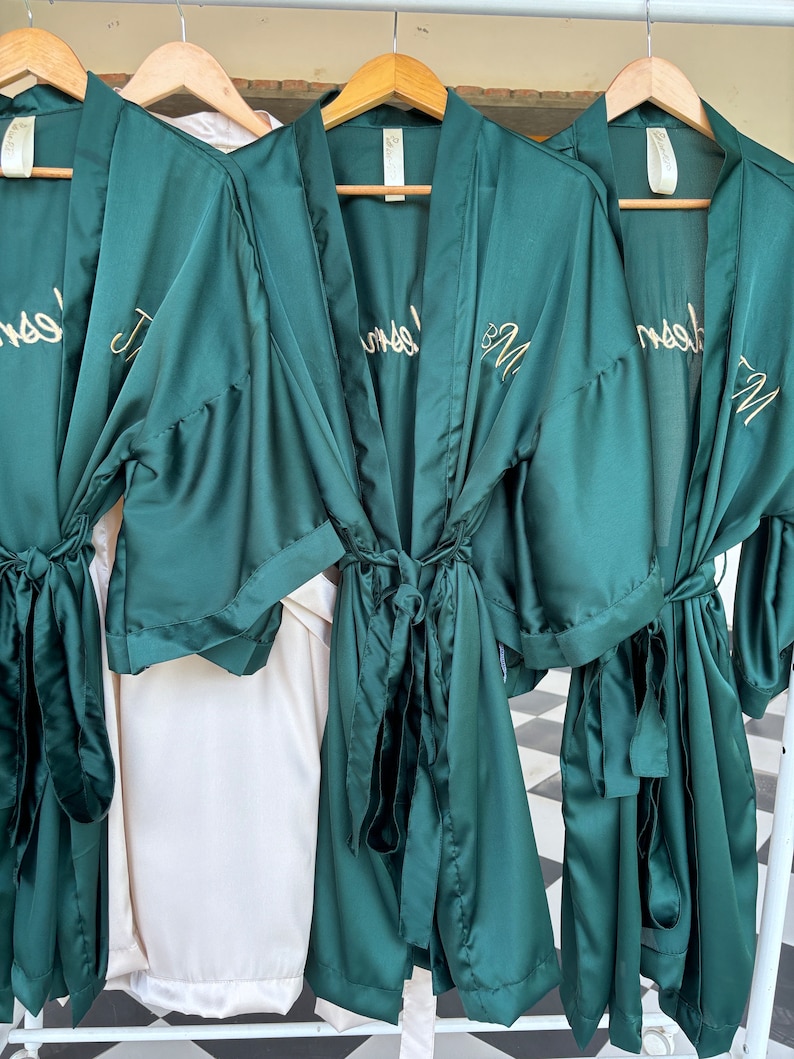 May include: Three green satin robes with gold embroidery. The robes have a sash tie and are hanging on wooden hangers. The embroidery on the robes reads "Bride" and "Bridesmaid".