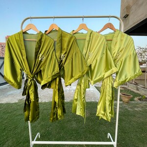 K&ouml;nnte beinhalten: Vier olivgr&uuml;ne Satin-Badem&auml;ntel h&auml;ngen an einem wei&szlig;en Gestell. Jeder Bademantel hat einen passenden Bindeg&uuml;rtel und gestickte Initialen. Die Badem&auml;ntel werden im Freien mit einem klaren blauen Himmel im Hintergrund ausgestellt.