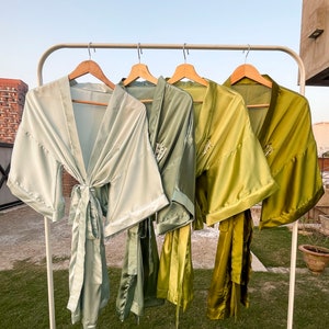 Op de afbeelding: Vier satijnen gewaden in verschillende tinten groen hangen aan een witte rek. De gewaden zijn allemaal in de taille gebonden en hebben lange mouwen.