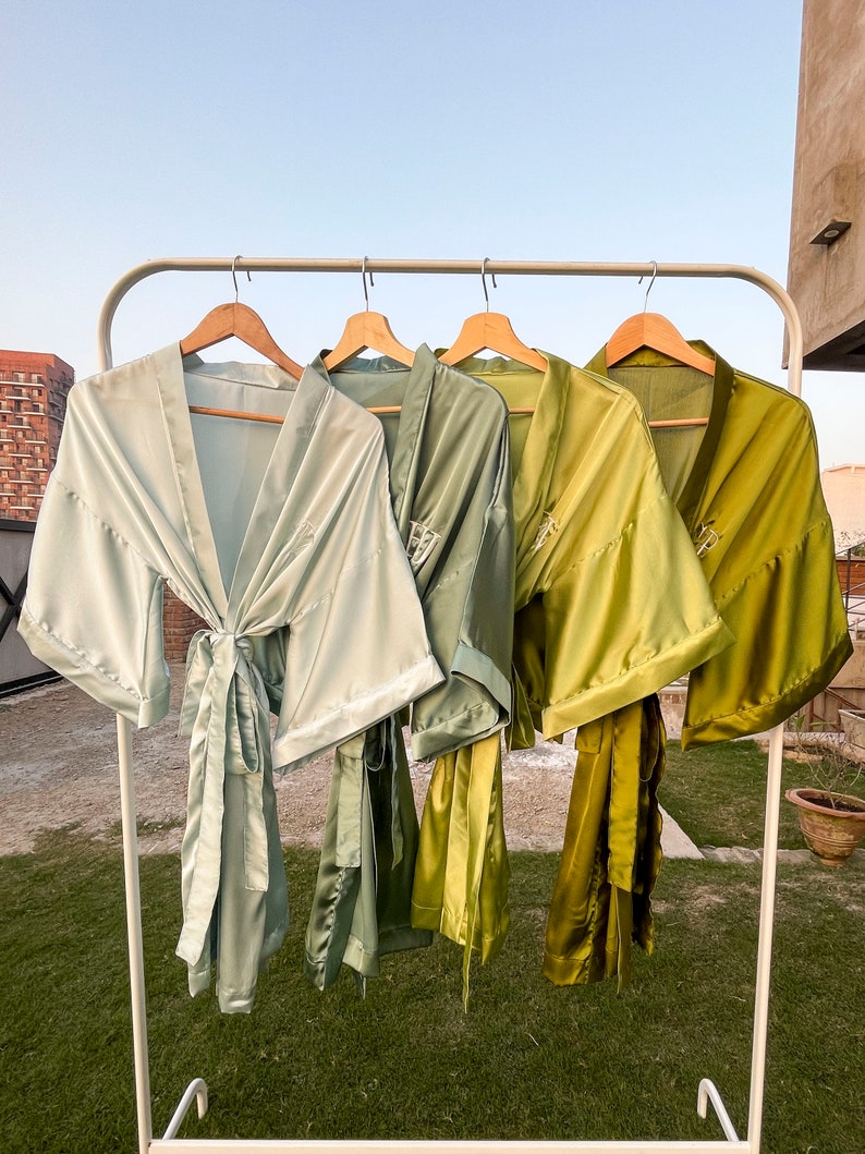 Op de afbeelding: Vier satijnen gewaden in verschillende tinten groen hangen aan een witte rek. De gewaden zijn allemaal in de taille gebonden en hebben lange mouwen.