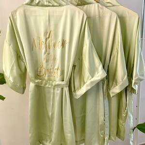 May include: Three light green satin robes with gold lettering on the back. The robe on the left says "Mother of the Bride".