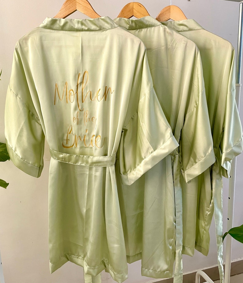 May include: Three light green satin robes with gold lettering on the back. The robe on the left says "Mother of the Bride".