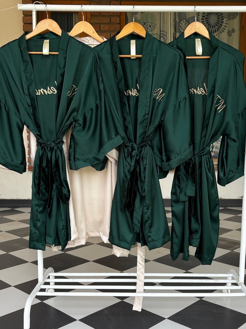 May include: Three dark green satin robes with gold embroidery hanging on a white rack. The robes have the names "Jess", "Emily", and "Jenna" embroidered on them.