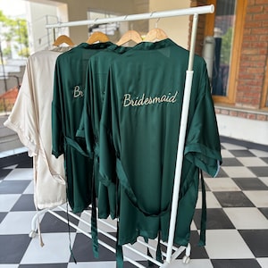 May include: Three green satin robes with gold embroidery hanging on a white rack. The robe on the right has the word "bridesmaid" embroidered on the back.