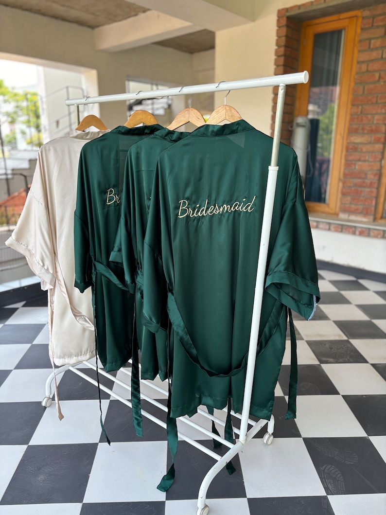 May include: Three green satin robes with gold embroidery hanging on a white rack. The robe on the right has the word "bridesmaid" embroidered on the back.