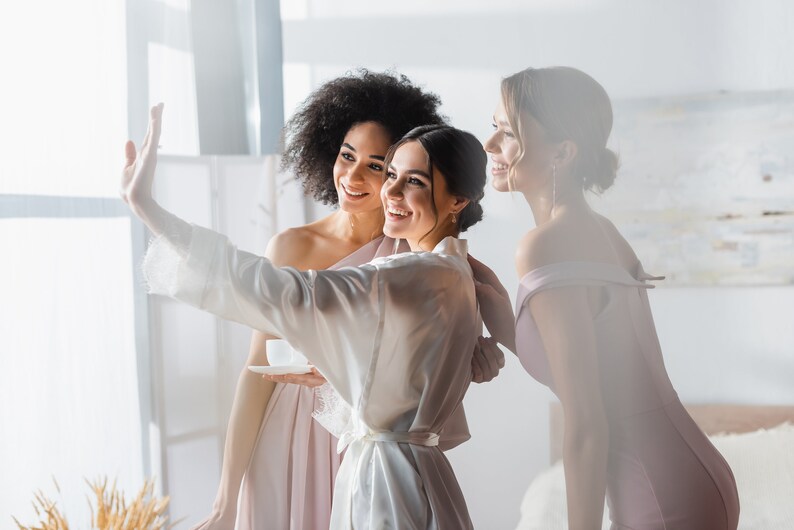 May include: Three women in a room, two wearing pink dresses and one wearing a white robe, smiling and taking a selfie.