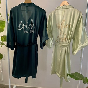 May include: Two satin robes hanging on a white rack. The robe on the left is dark green with the text "Bride Est. 2022" in gold glitter. The robe on the right is light green with the text "Mother of the Bride" in gold glitter.