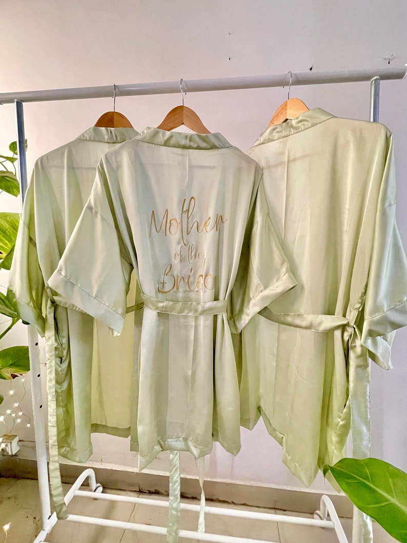 May include: Three light green satin robes hanging on a rack. The middle robe has the text "Mother of the Bride" embroidered on the back in gold lettering.