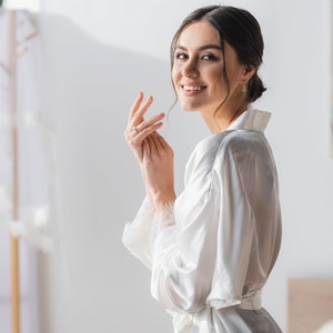 May include: A woman in a white satin robe with a lace trim, smiling and looking over her shoulder. She is wearing a ring on her finger.