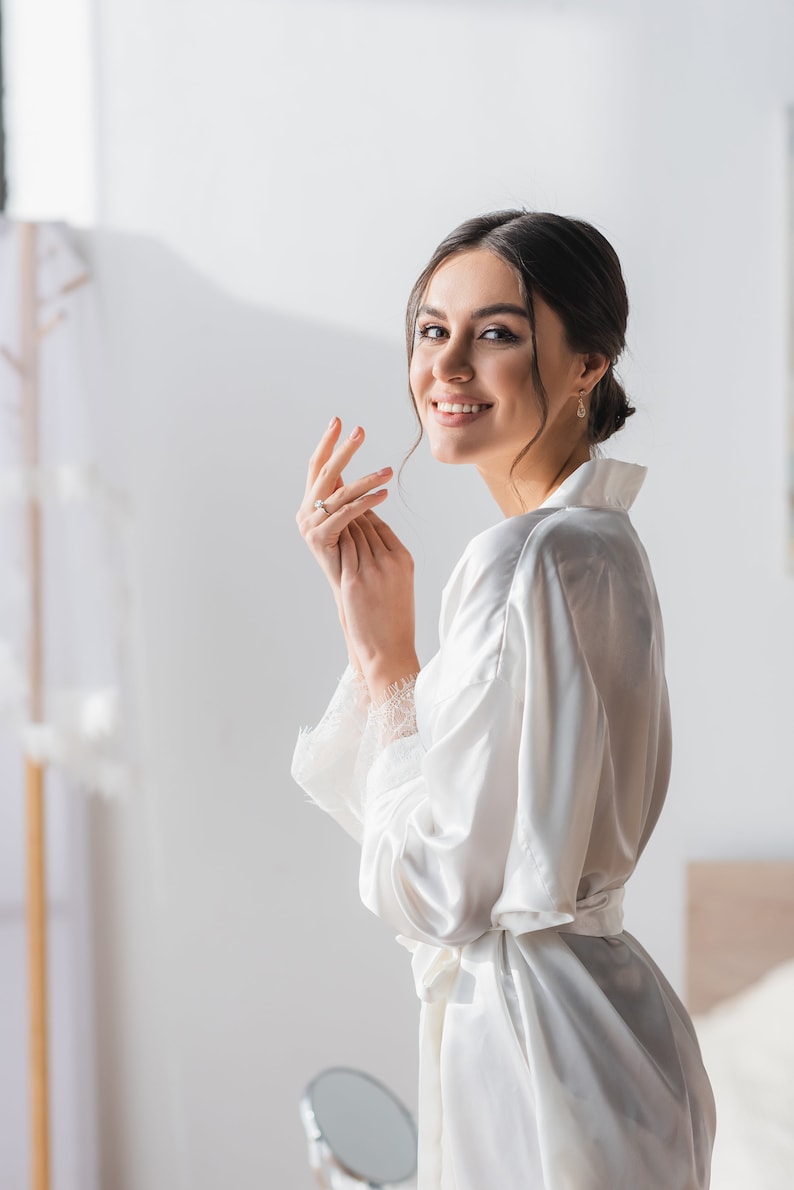 May include: A woman in a white satin robe with a lace trim, smiling and looking over her shoulder. She is wearing a ring on her finger.