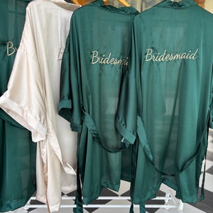 May include: Three satin robes hanging on a rack. Two robes are dark green and one is light beige. The robes have the word "bridesmaid" embroidered on the back in gold thread.