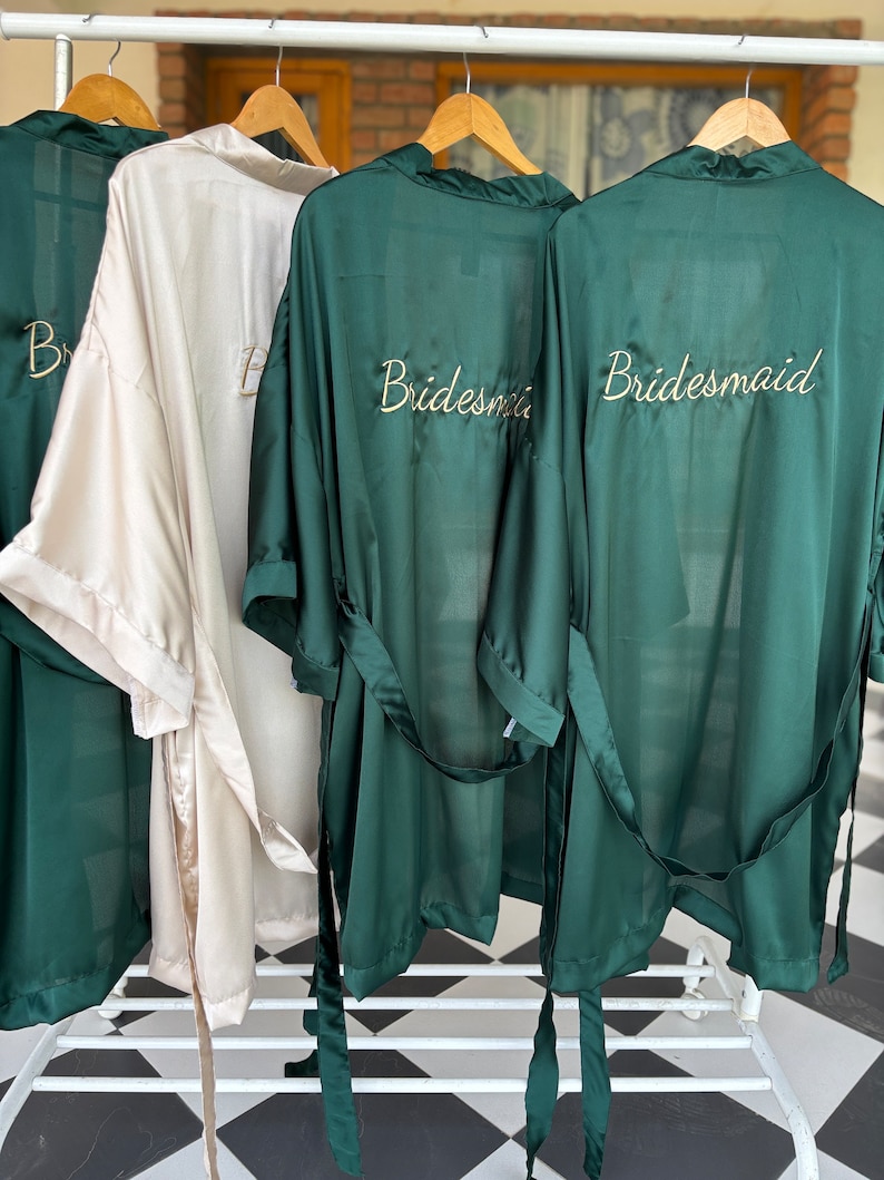 May include: Three satin robes hanging on a rack. Two robes are dark green and one is light beige. The robes have the word "bridesmaid" embroidered on the back in gold thread.