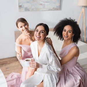 May include: Three women in a bedroom, two wearing pink dresses and one wearing a white robe, smiling at the camera. The woman in the robe is holding a cup of coffee.