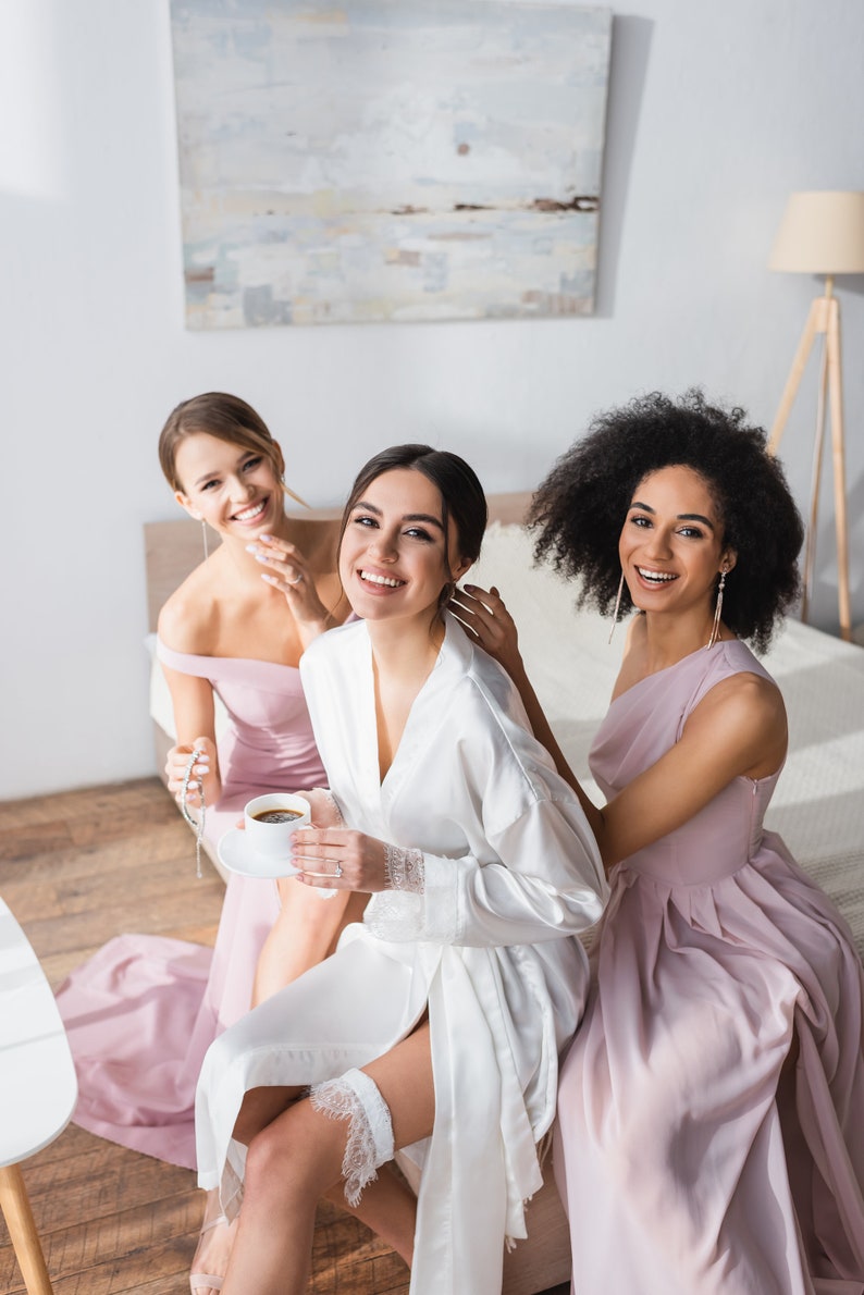 May include: Three women in a bedroom, two wearing pink dresses and one wearing a white robe, smiling at the camera. The woman in the robe is holding a cup of coffee.
