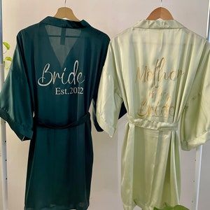May include: Two satin robes, one green and one light green, hanging on a white rack. The green robe has "Bride Est. 2012" embroidered on the back. The light green robe has "Mother of the Bride" embroidered on the back.