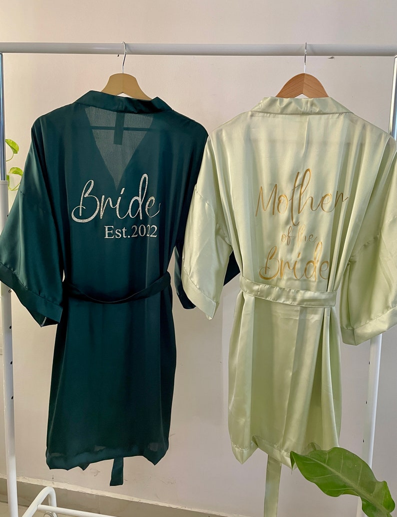 May include: Two satin robes, one green and one light green, hanging on a white rack. The green robe has "Bride Est. 2012" embroidered on the back. The light green robe has "Mother of the Bride" embroidered on the back.