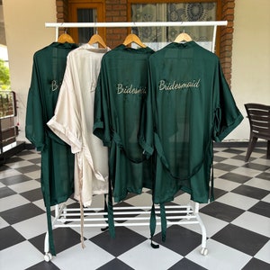 May include: Four satin robes hanging on a rack. Three robes are dark green and one is cream. The green robes have the word "Bride" or "Bridesmaid" embroidered on the back in gold thread.